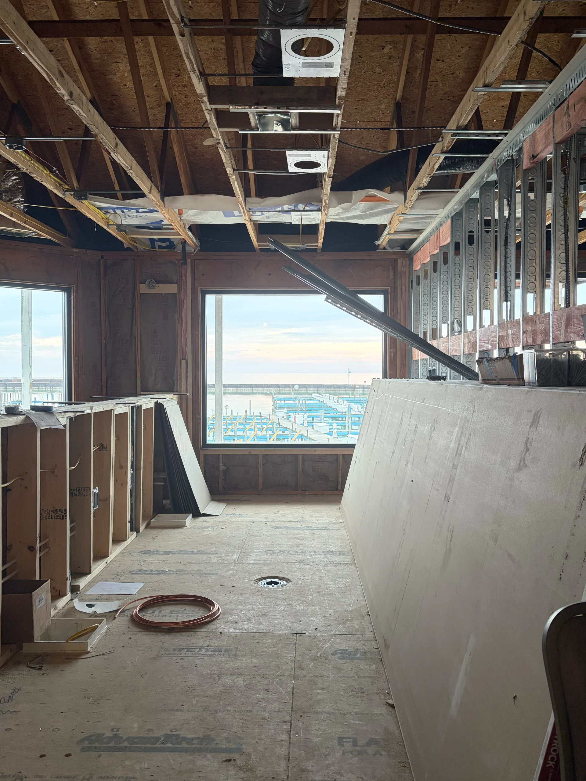 The interior of a waterfront building under construction, showing wood framing for a bar and exposed ceiling joists. The windows frame a view of a marina with boat docks.
