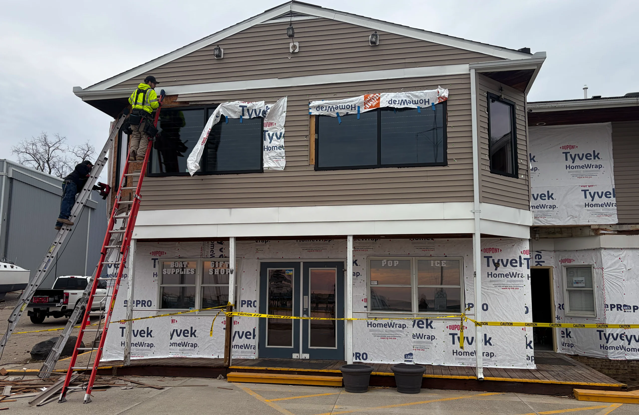 Two workers on ladders install windows and siding on a tan, two-story building undergoing renovation, partially covered in white Tyvek HomeWrap.