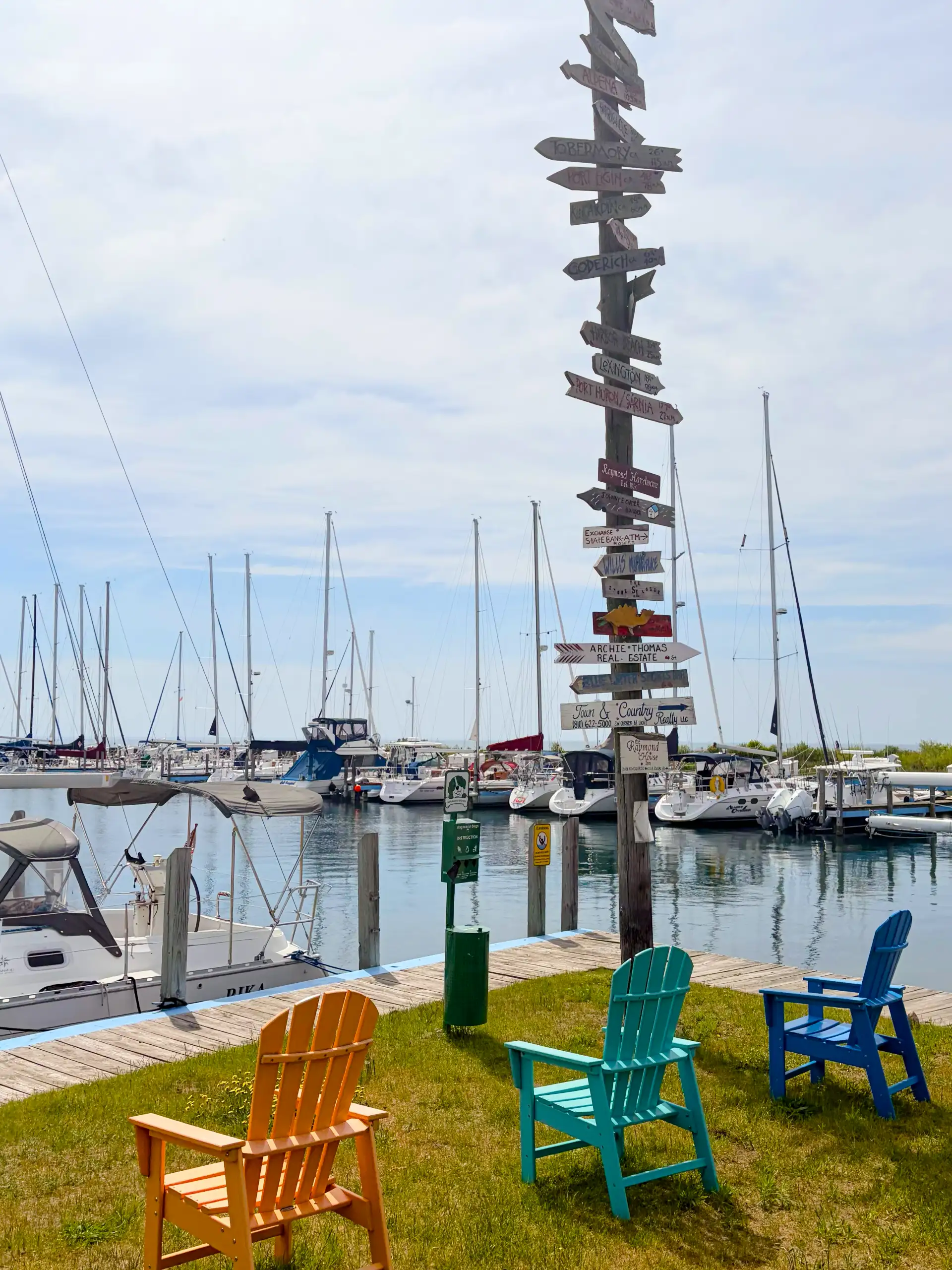 A tall wooden post covered in directional signs overlooks a sunny marina. Three bright orange, teal, and blue Adirondack chairs sit on the grassy dock next to many moored sailboats.