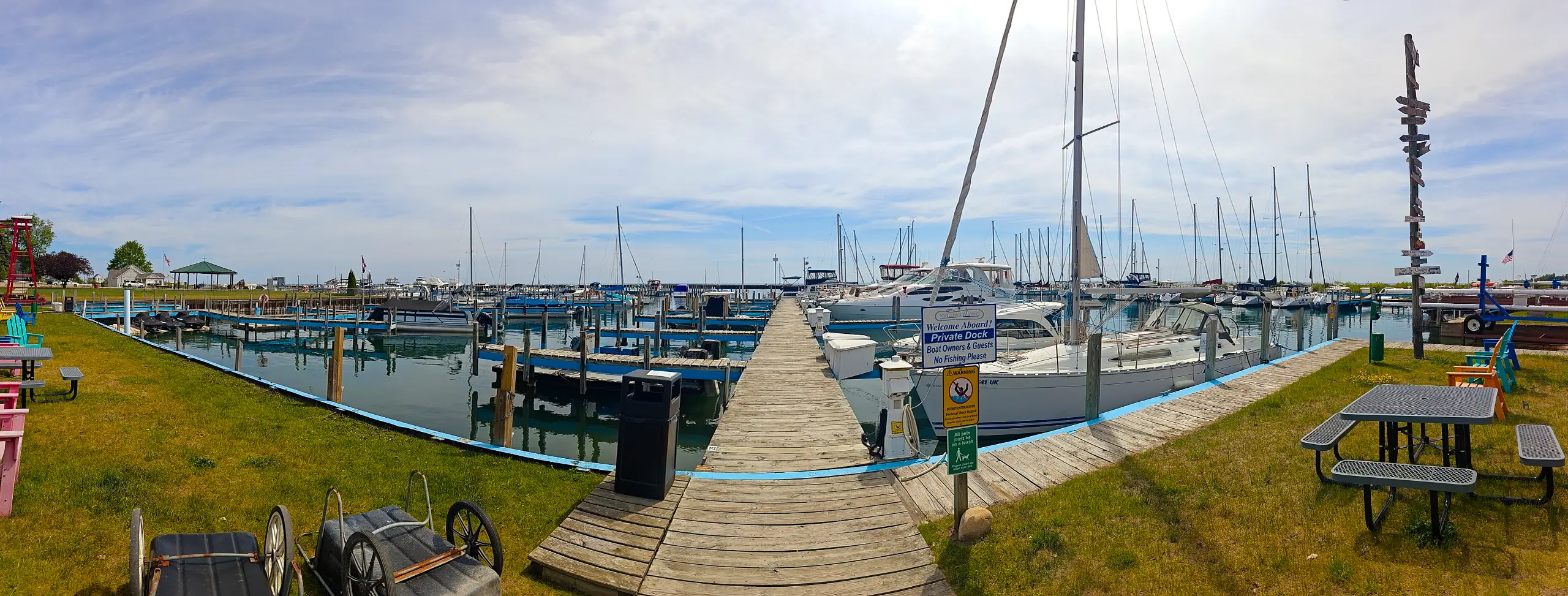 A panoramic image of a sunny marina featuring a wooden dock leading into the water, surrounded by numerous sailboats and powerboats.