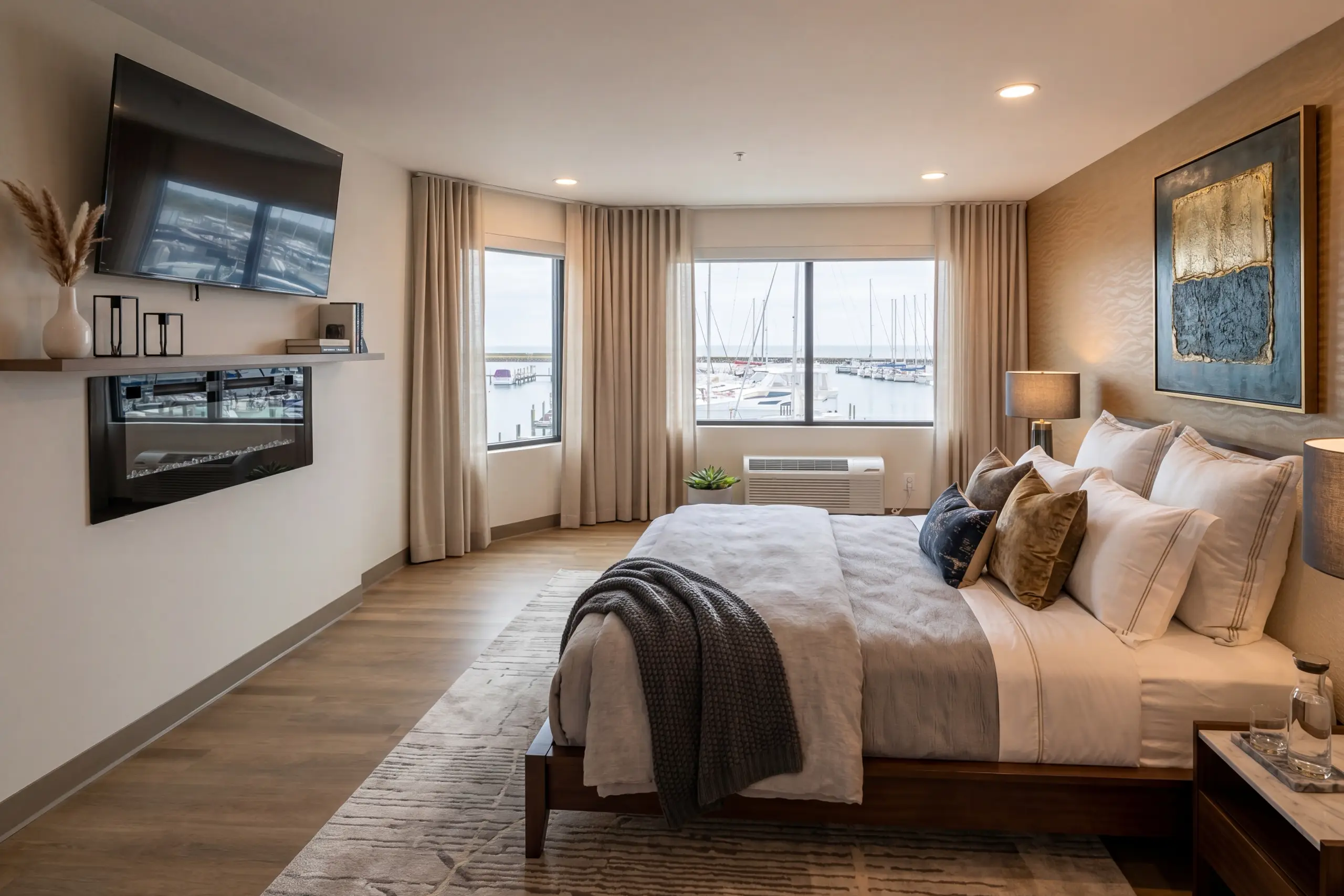 A large bed sits in a modern hotel room with a TV and fireplace, facing windows overlooking a boat-filled marina.