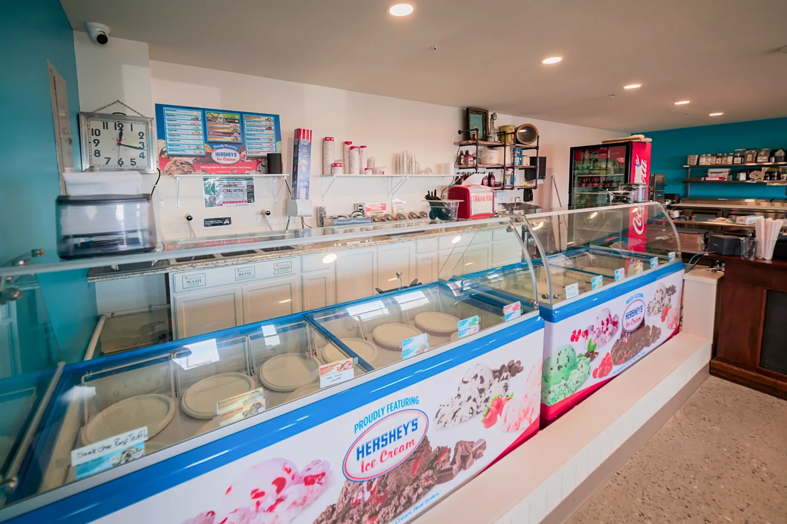 A brightly lit ice cream parlor counter featuring a long glass freezer display case branded with Hershey's Ice Cream, set against teal and white walls.