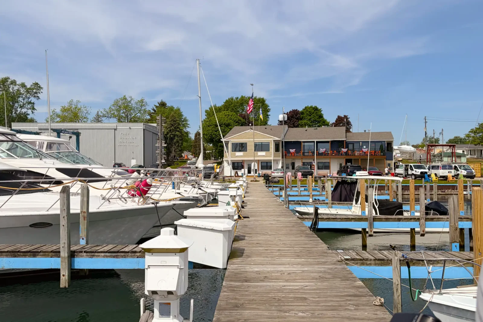 A long wooden dock extends between moored motorboats and sailboats toward a multi-level tan and blue marina building on a sunny day.