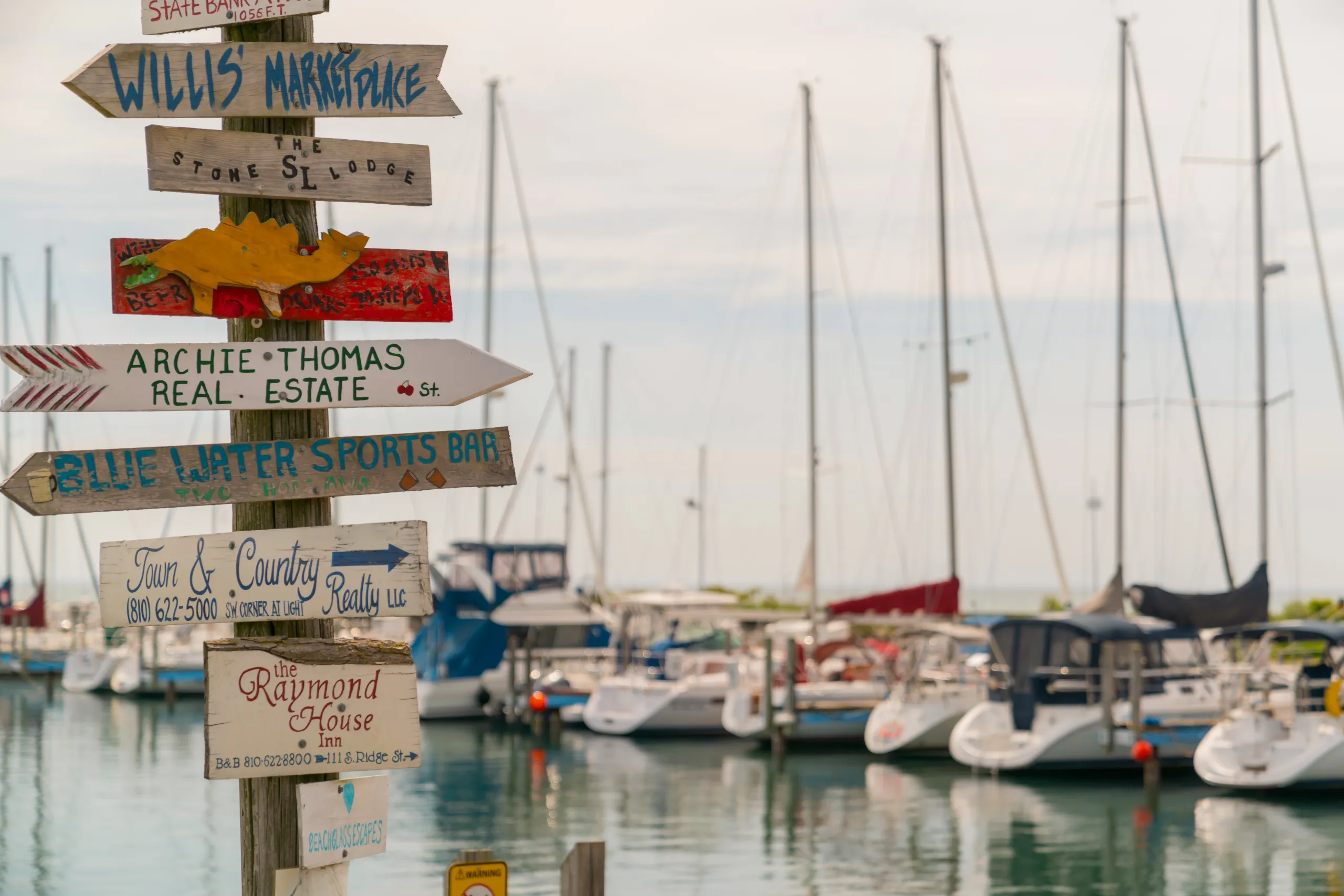 A post holding colorful directional signs for various local businesses stands at a sunny marina filled with the masts of docked sailboats.
