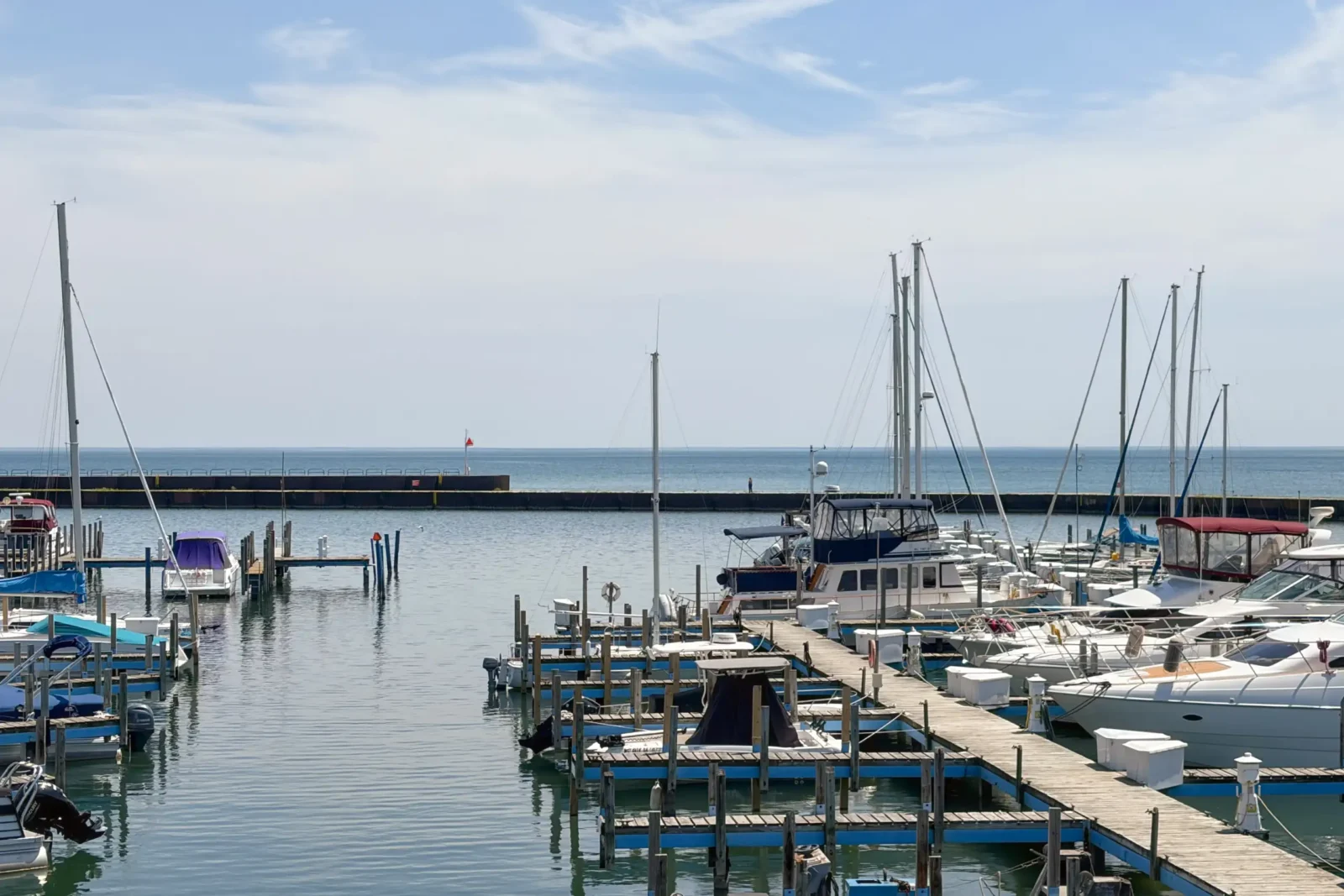 A view across a large, calm marina filled with powerboats and sailboats docked at wooden piers. A dark breakwater wall separates the harbor from the open blue water.