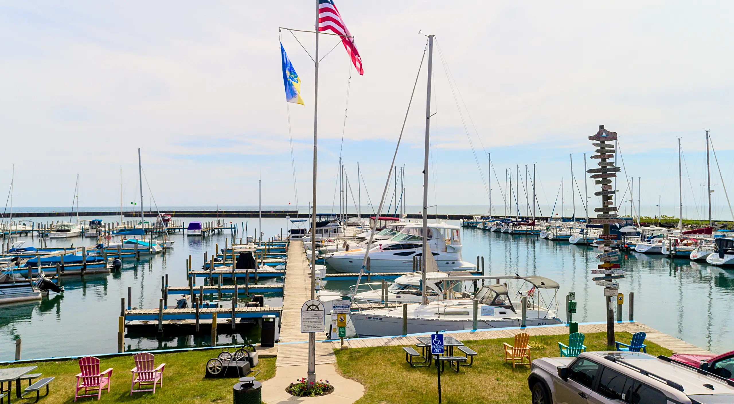 A marina view on a bright day with many sailboats and motorboats tied up along the wooden docks. The foreground features a grassy area with picnic tables and colorful seating.