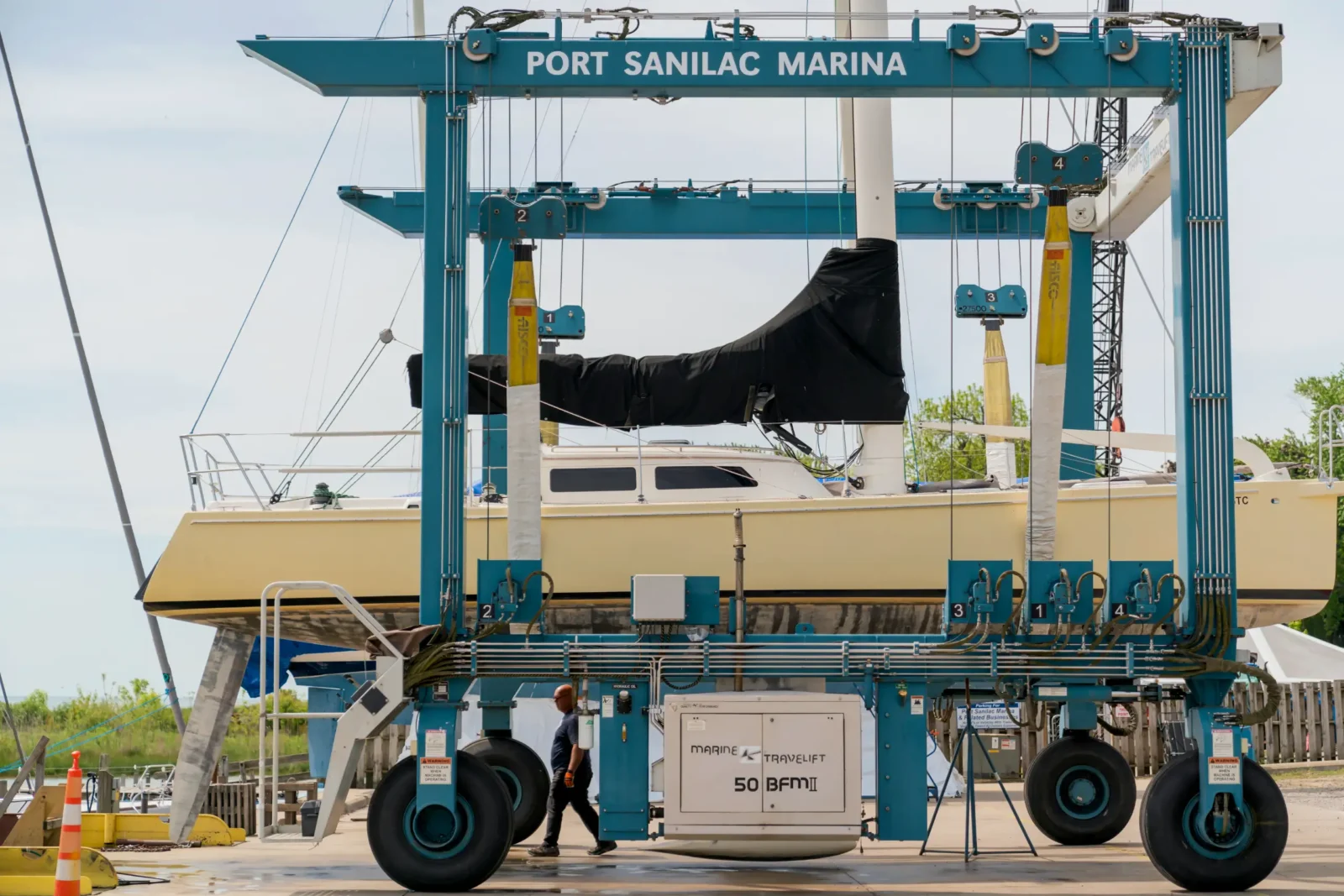 A large blue Marine Travelift hoist at Port Sanilac Marina holds a yellow sailboat. A marina worker walks past the lift's wheels.