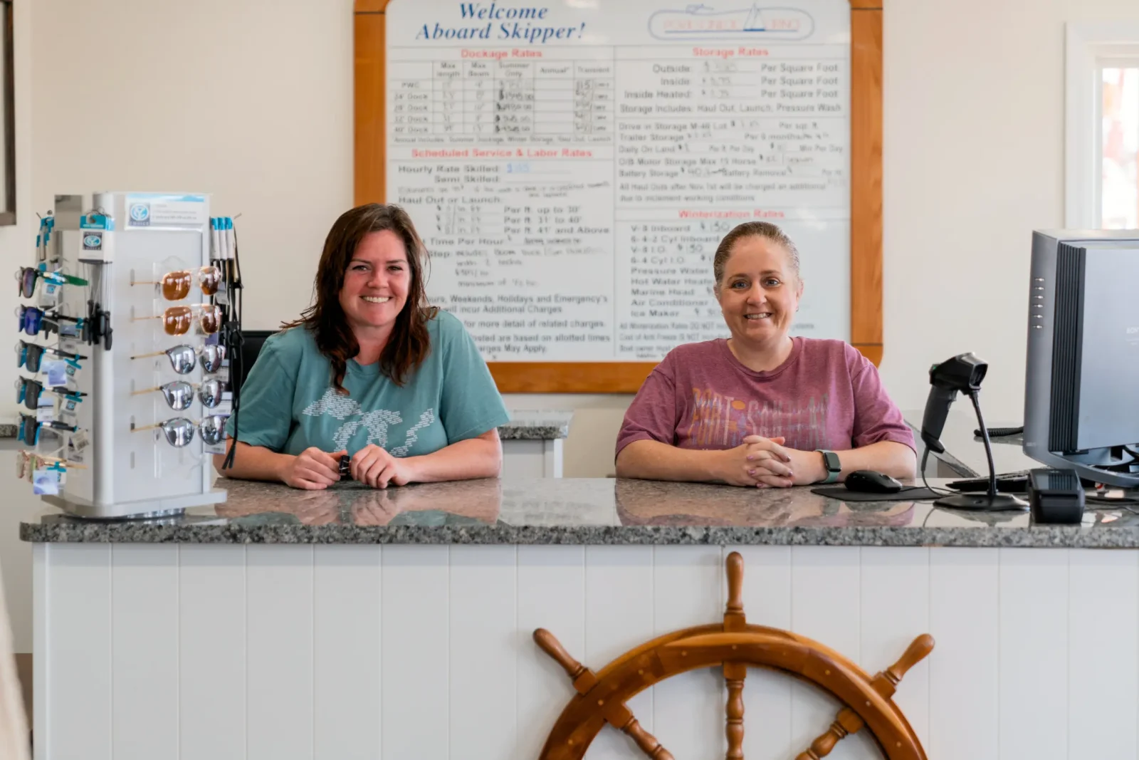 Two smiling women, marina staff, stand behind a counter featuring a prominent ship's wheel decoration and a rate board detailing dockage and storage fees.