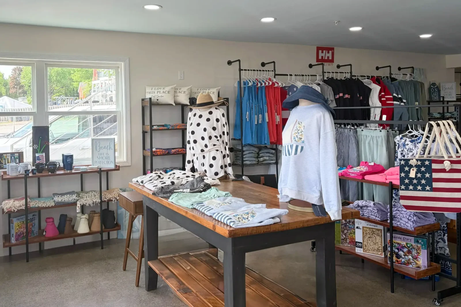 Retail display inside a marina shop with racks of beach apparel and casual clothing, a central table, and an American flag tote bag.