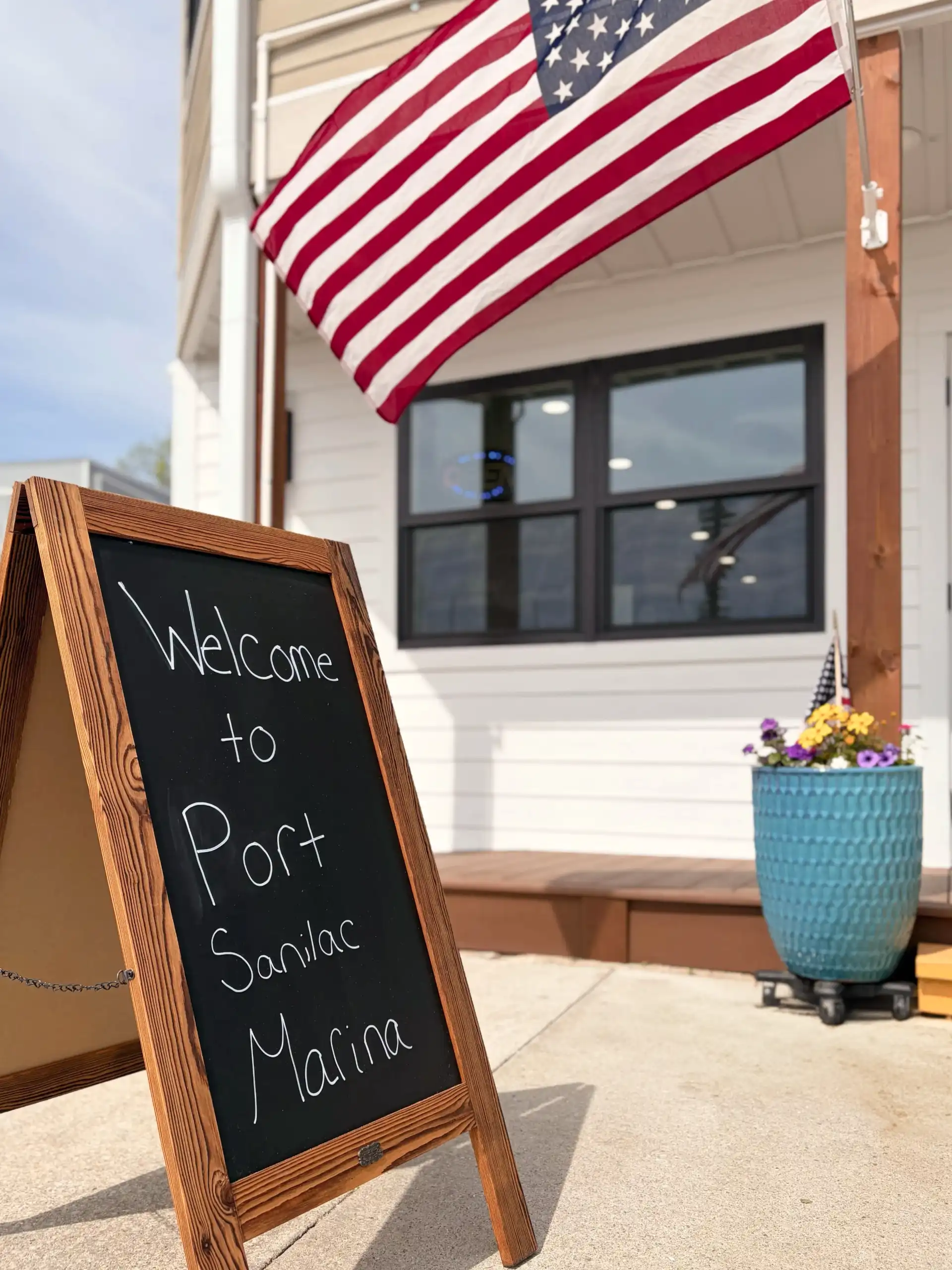 A wooden A-frame chalkboard sign reads "Welcome to Port Sanilac Marina" on a sunny day, with a large American flag flying overhead.