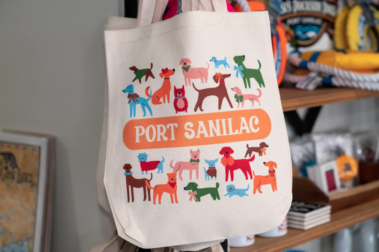 A canvas tote bag is printed with a cheerful collection of colorful dogs, featuring various breeds and accessories. The text "PORT SANILAC" is centered.