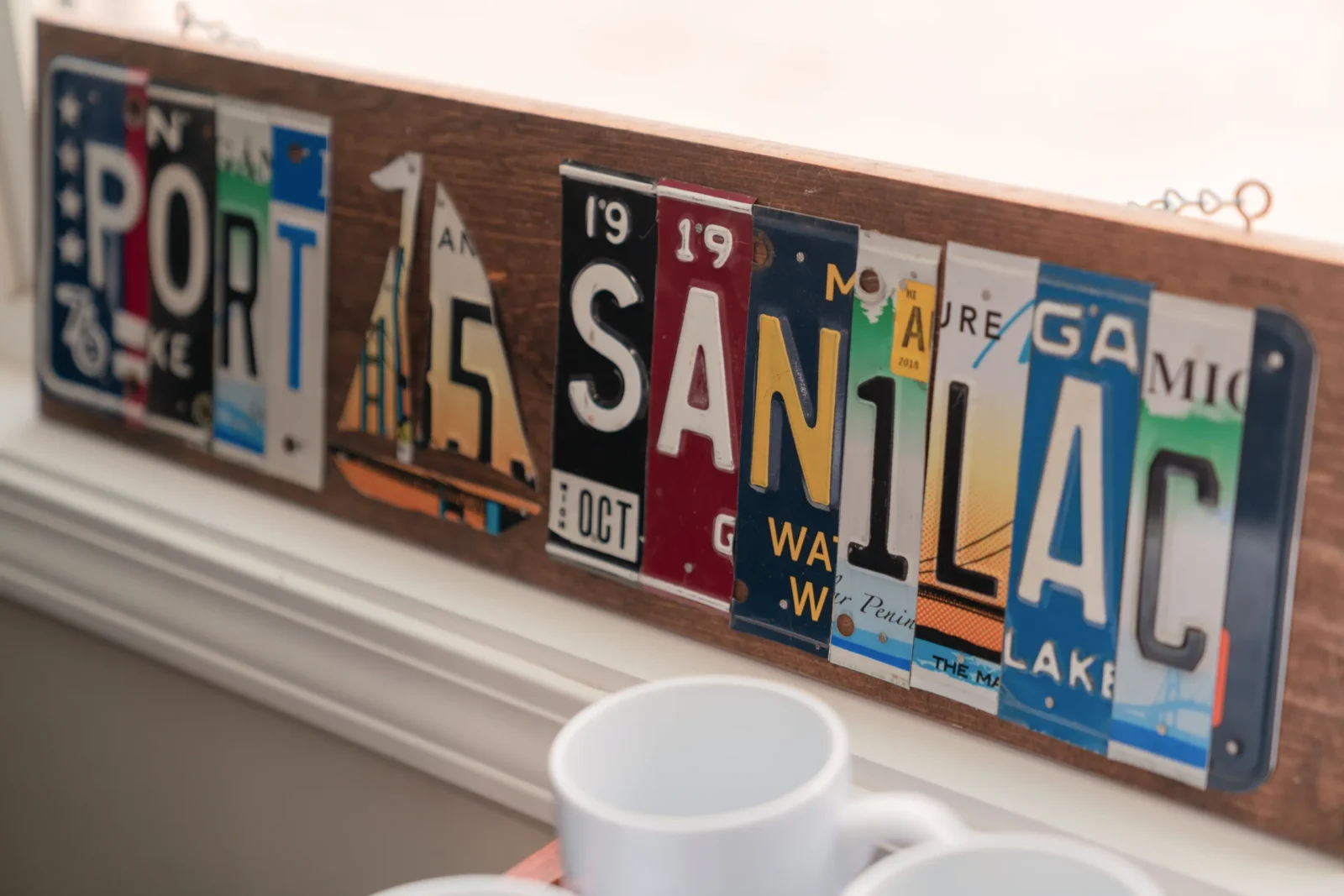A decorative sign spelling "PORT SANILAC" is crafted from repurposed license plates and mounted on a wooden board. A license plate sailboat separates the words.