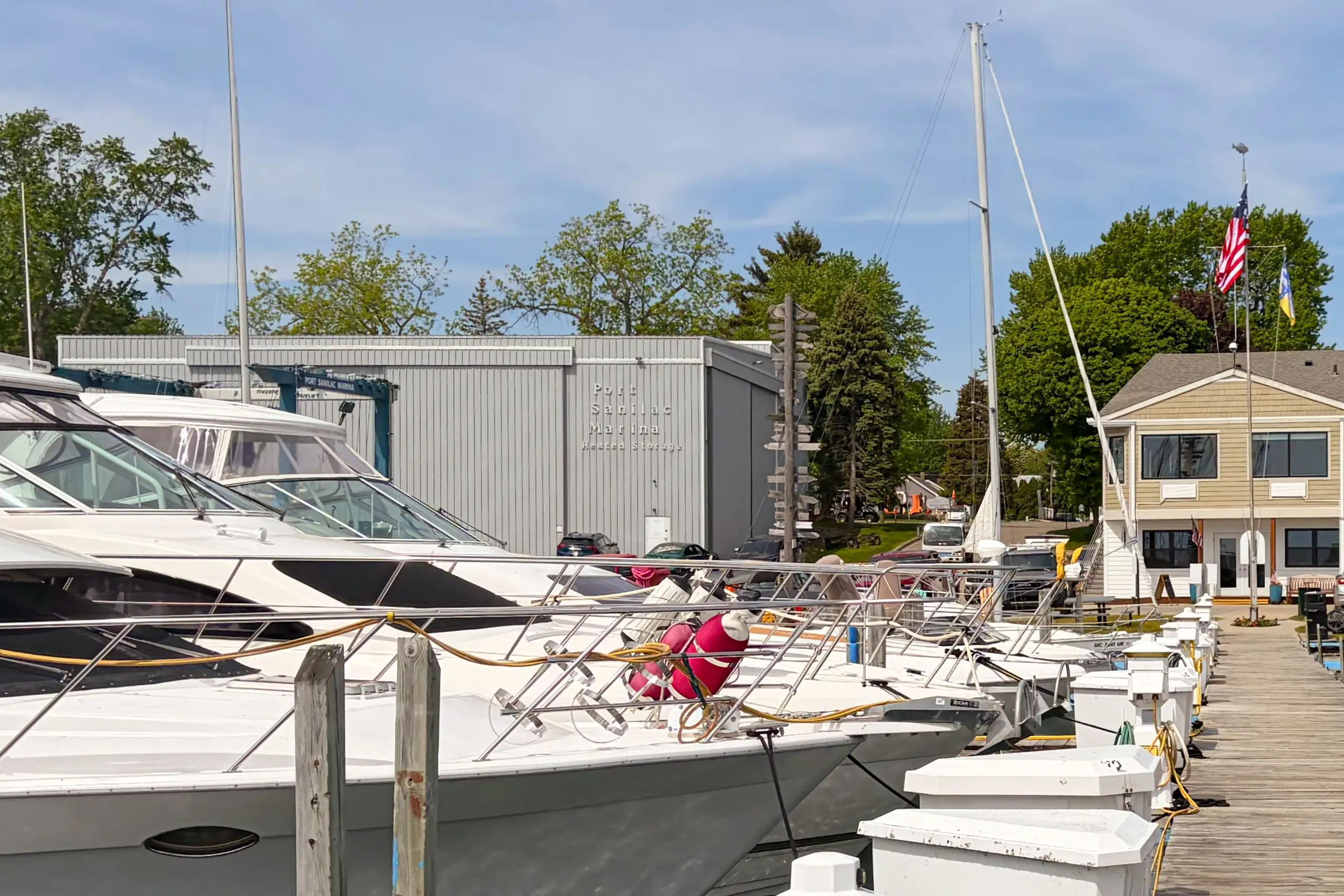 Yachts are docked along a wooden pier at Port Sanilac Marina, backed by a gray metal heated storage building and a small office building.