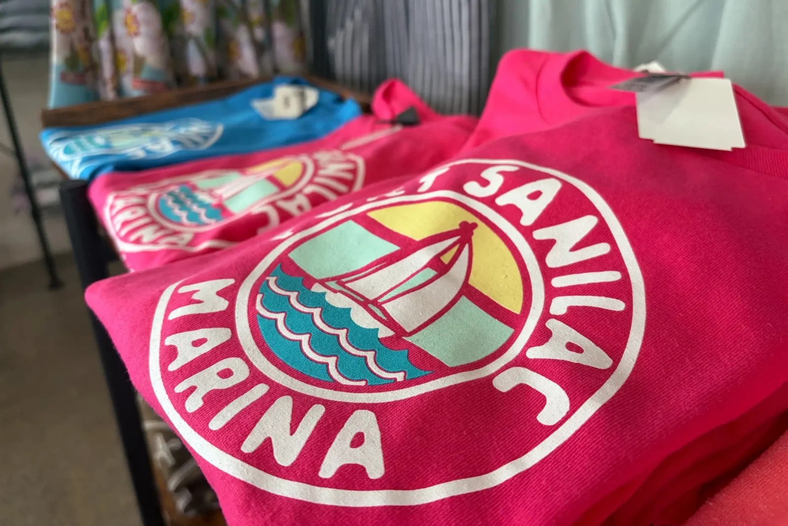 A stack of bright pink t-shirts with a circular Port Sanilac Marina logo showing a sailboat on waves.