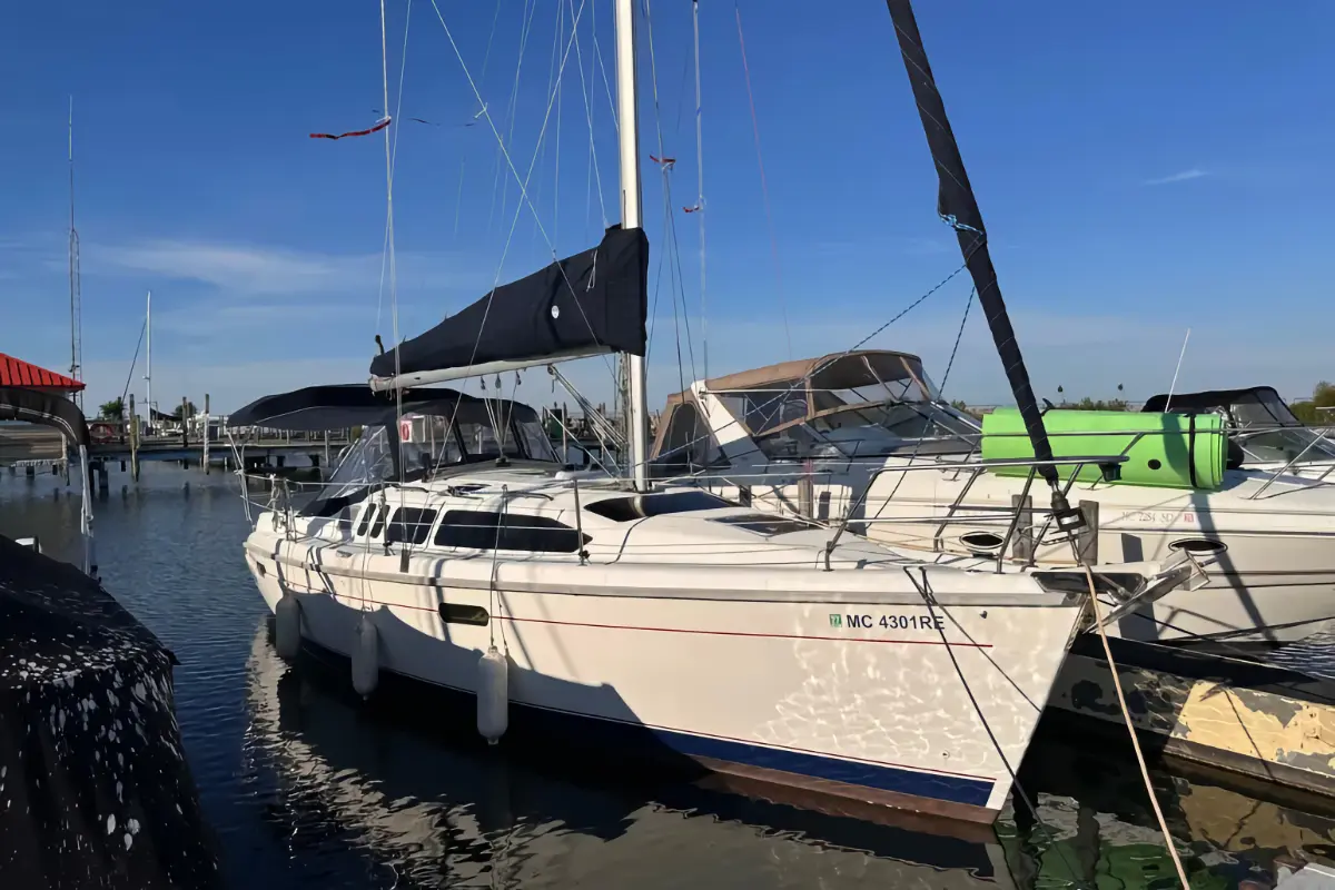 A sleek white sailboat is moored at a dock in a sunny marina, featuring dark blue canvas covers and secured with fenders.