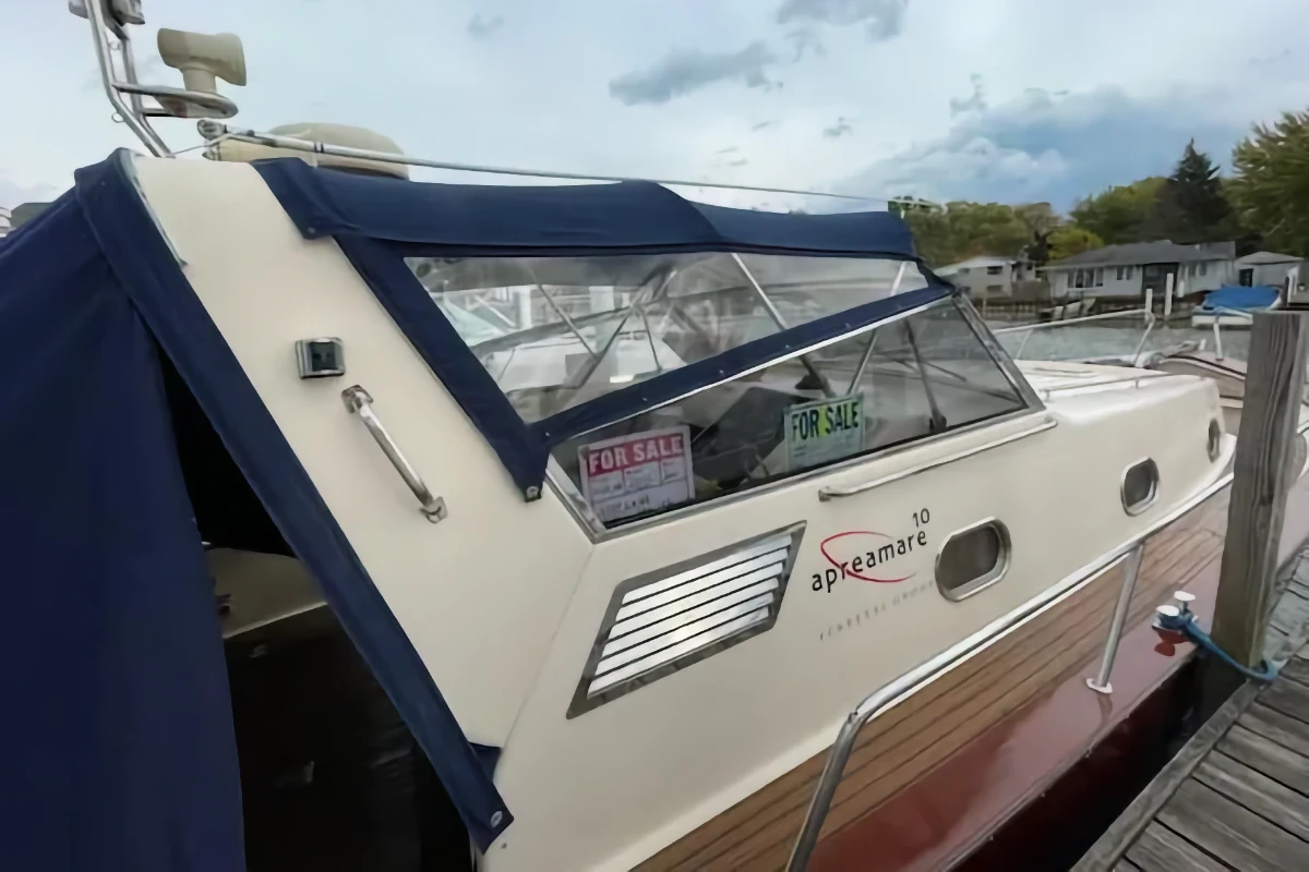 A beige Apreamare cabin cruiser with a navy blue canvas enclosure is secured to a wooden dock. Two "For Sale" signs are visible behind the windshield.