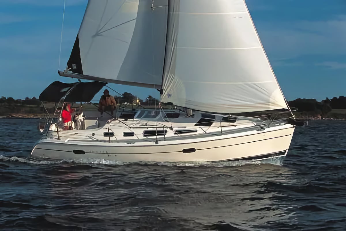 A large white sailboat with two passengers in the cockpit cruises on dark water under a bright blue sky, powered by its full white sails.