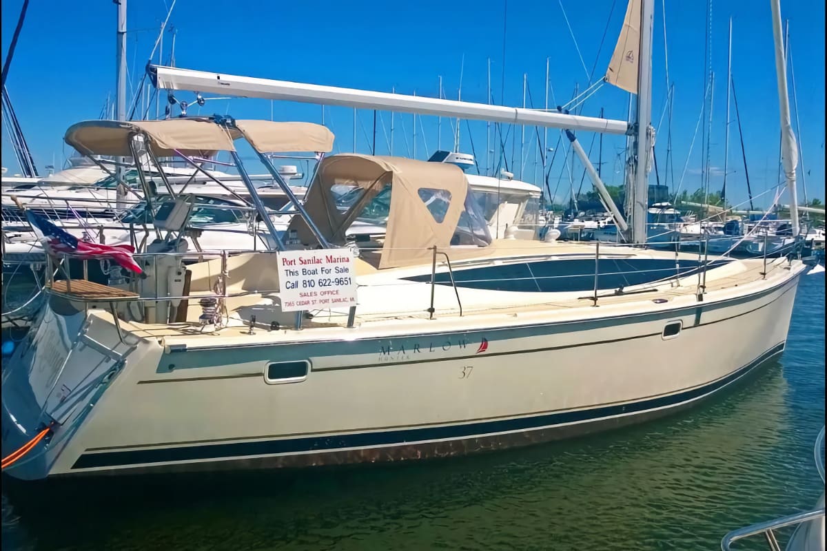A beige Marlow Hunter 37 sailboat is docked in a sunny marina, featuring tan cockpit covers, a blue hull stripe, and a 'For Sale' sign.
