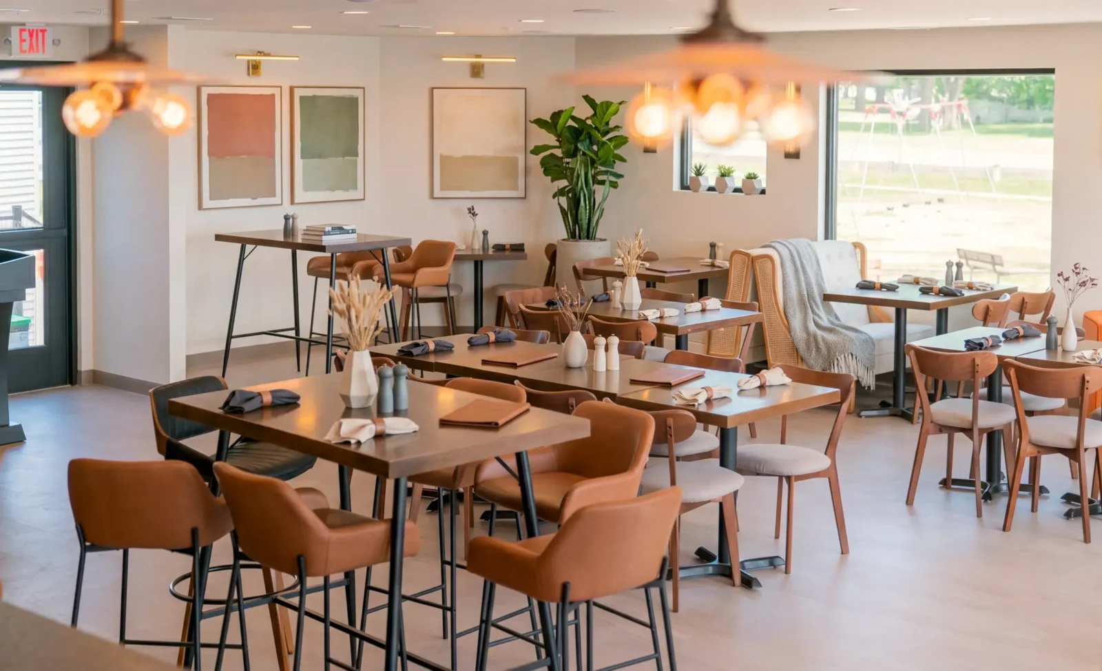 A bright, modern restaurant dining room features dark wood tables set for dining, surrounded by leather and wood chairs. Abstract art and large potted plants decorate the inviting space.