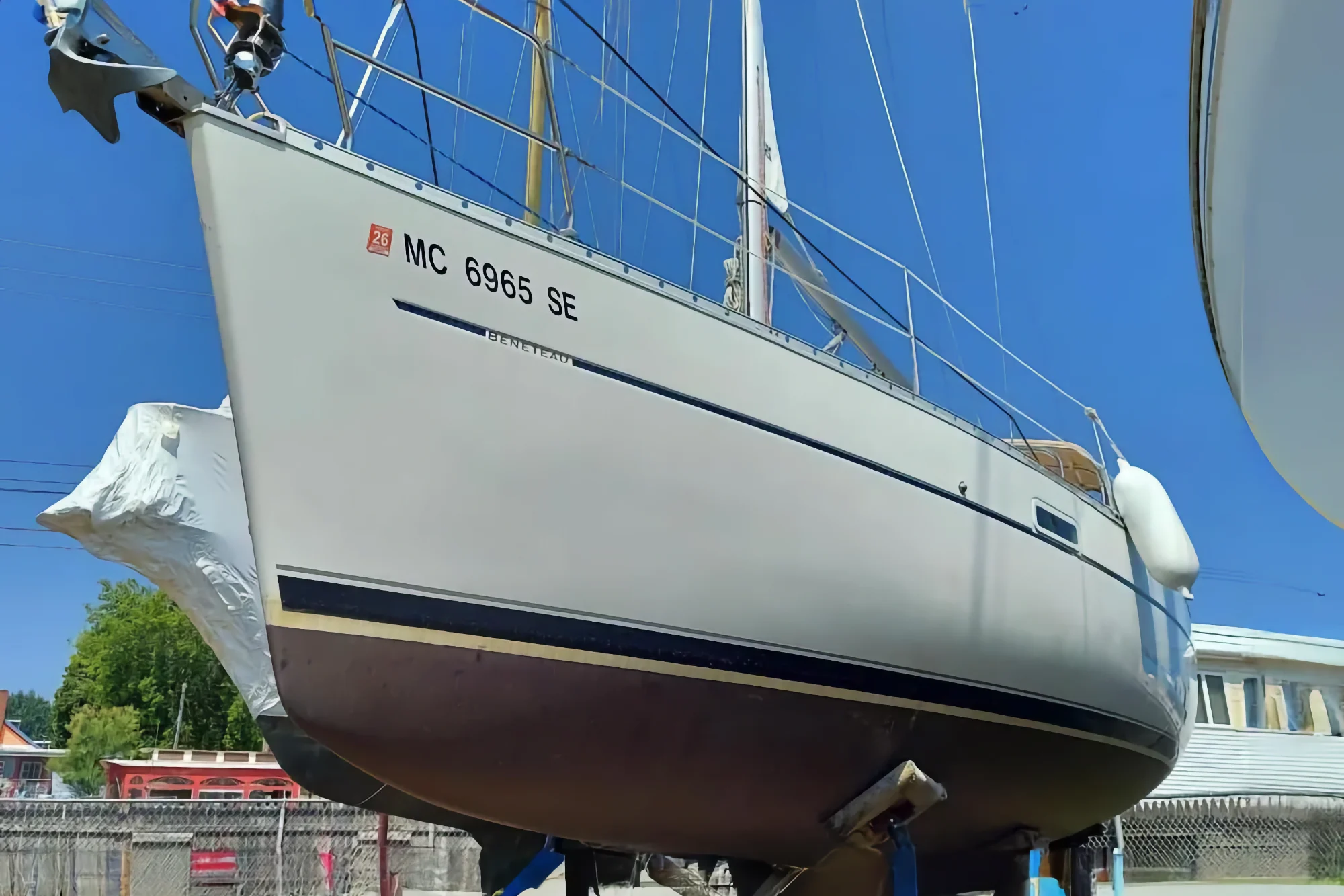 A white Beneteau 331 sailboat is out of the water, resting on stands in a sunny marina yard. The bow features the registration number MC 6965 SE.