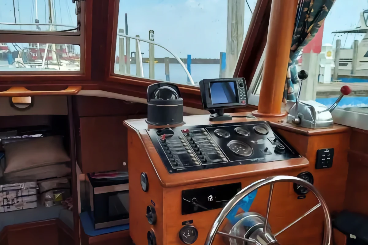 The interior helm station of a wooden boat cabin, featuring a steering wheel, navigation controls, gauges, and a compass, with a marina visible through the windshield.