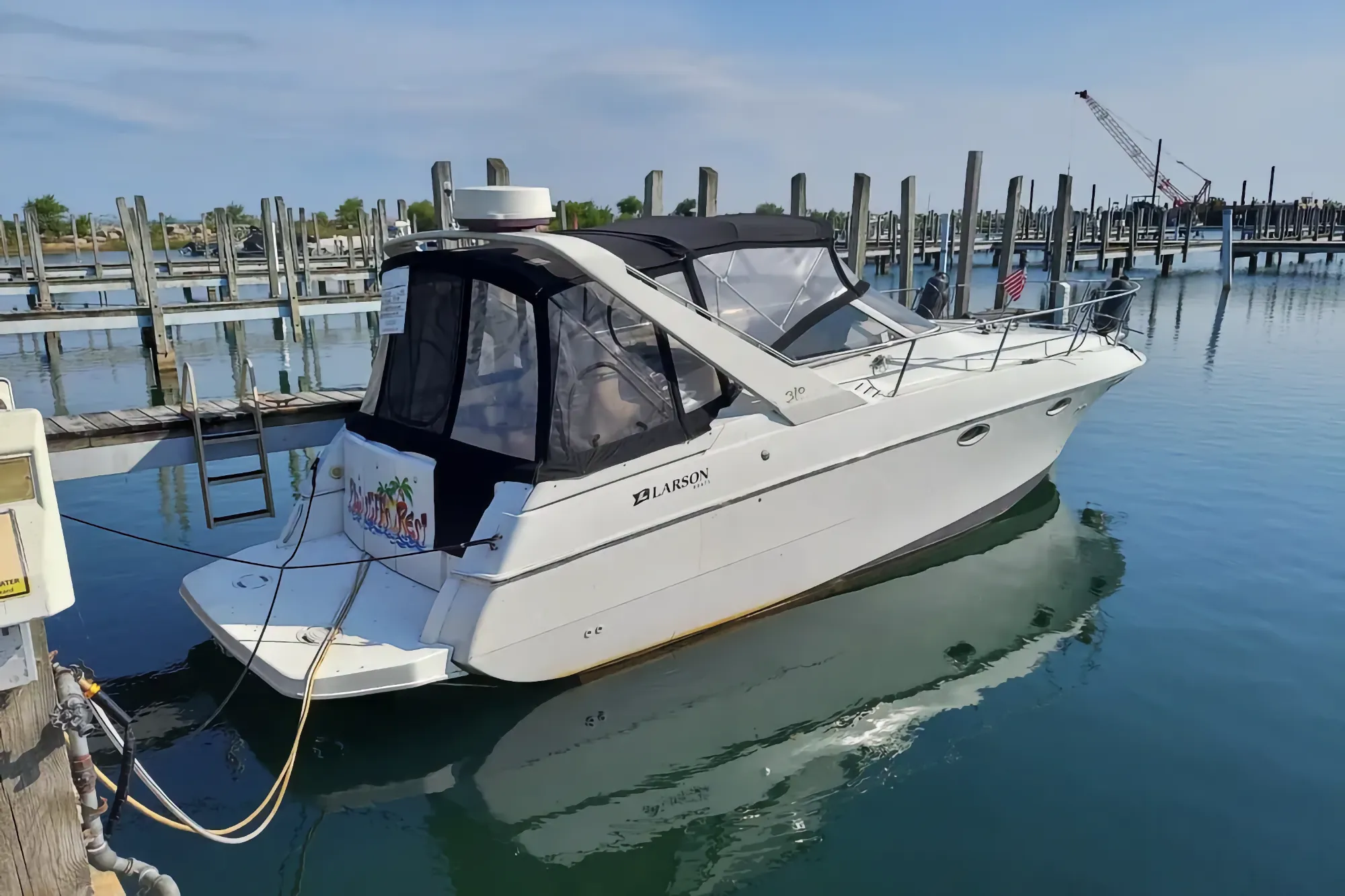 A white Larson Cabrio 310 cruiser with a black top floats peacefully, tied to a wooden dock at a marina on a sunny day.