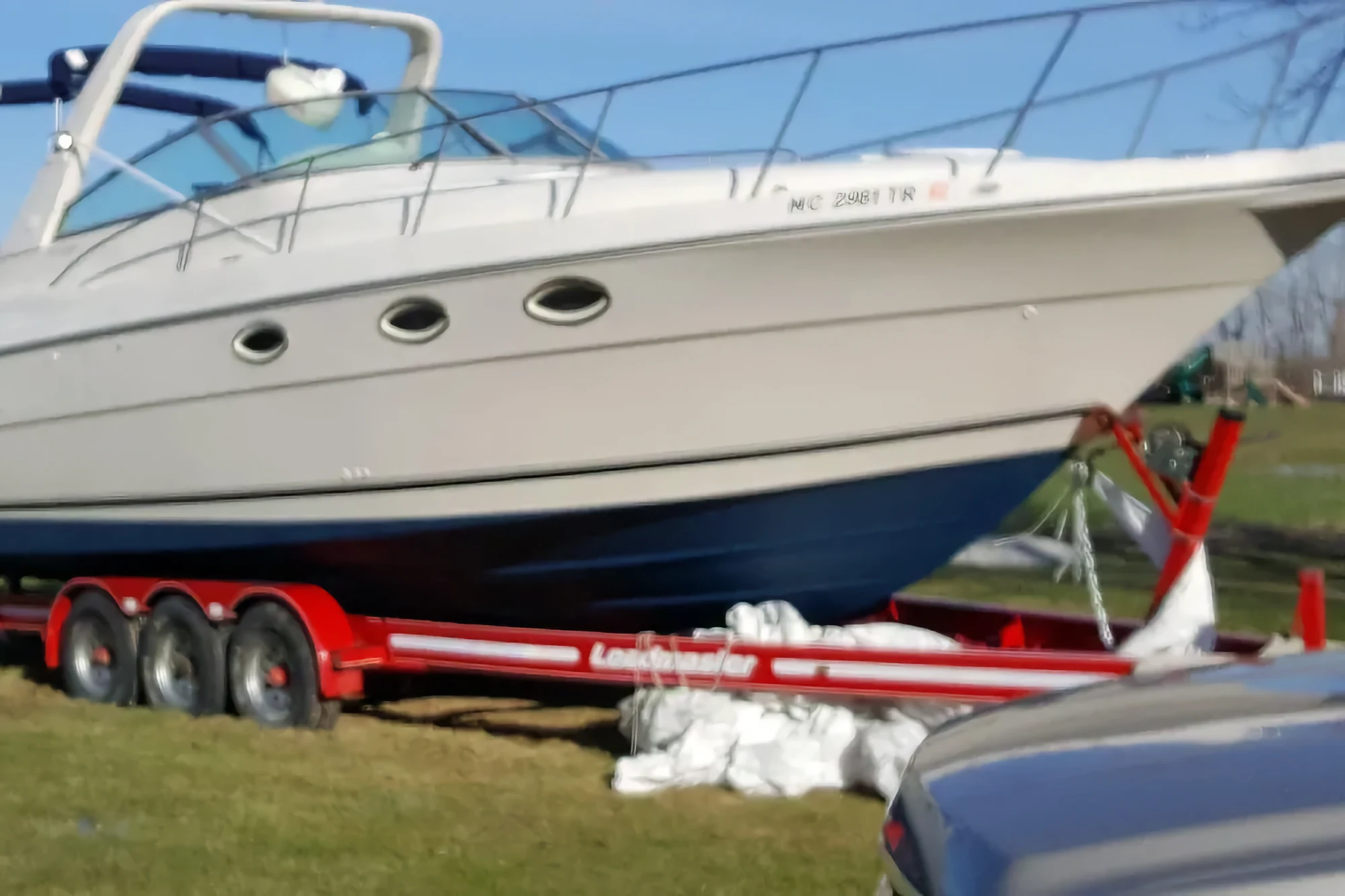 A large white and blue cruiser boat is displayed outside on a bright red triple-axle trailer under a clear blue sky.