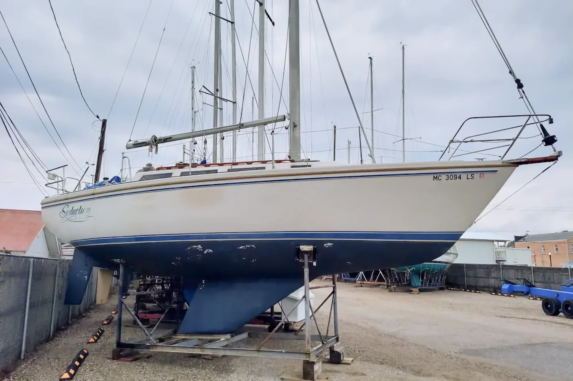 A white and blue sailboat named Seduction, with registration MC 3094 LS, sits on metal support stands in a dry dock yard under a cloudy sky. Other boat masts are visible in the background.